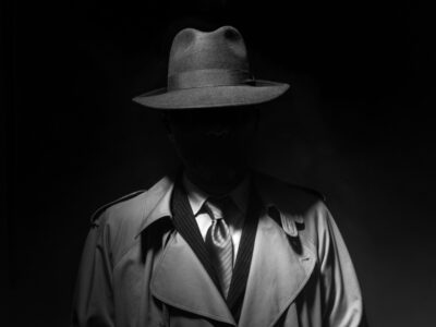 Man wearing a suit, trench coat and brimmed hat with face darkened.