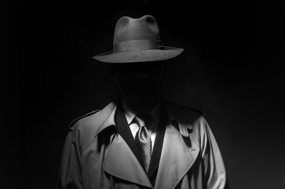 Man wearing a suit, trench coat and brimmed hat with face darkened.